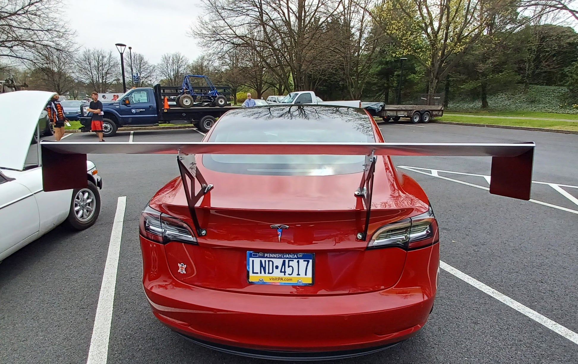 A red Tesla with a 9livesracing Tesla Model 3 Big Wang kit is parked in a lot, with its license plate "LND-4517" visible. The trunk is open and people are standing nearby. Trees and a grassy area are in the background.