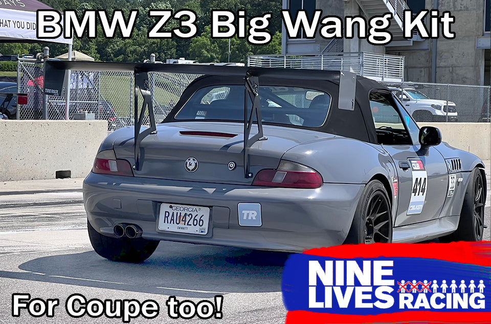 A silver BMW Z3 is shown on a racetrack parking area, featuring an impressive 9livesracing BMW Z3 Roadster Wing. The car has the number 44 on its side and an "RAU4266" license plate. Signs promoting "Nine Lives Racing" and the "BMW Z3 Roadster Wing - For Coupe Too!" boast about its improved lift-to-drag ratio.