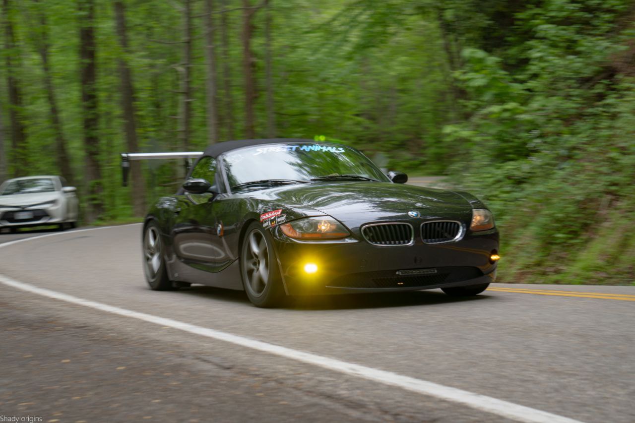 A black BMW Z4 sports car, equipped with a 9livesracing BMW z4 Big Wang kit (2003-08 E85), is driving through a winding forest road. The car has decals on the windshield and front bumper. Another vehicle is visible in the background on the same road, while the surrounding area is lush with green trees and foliage.