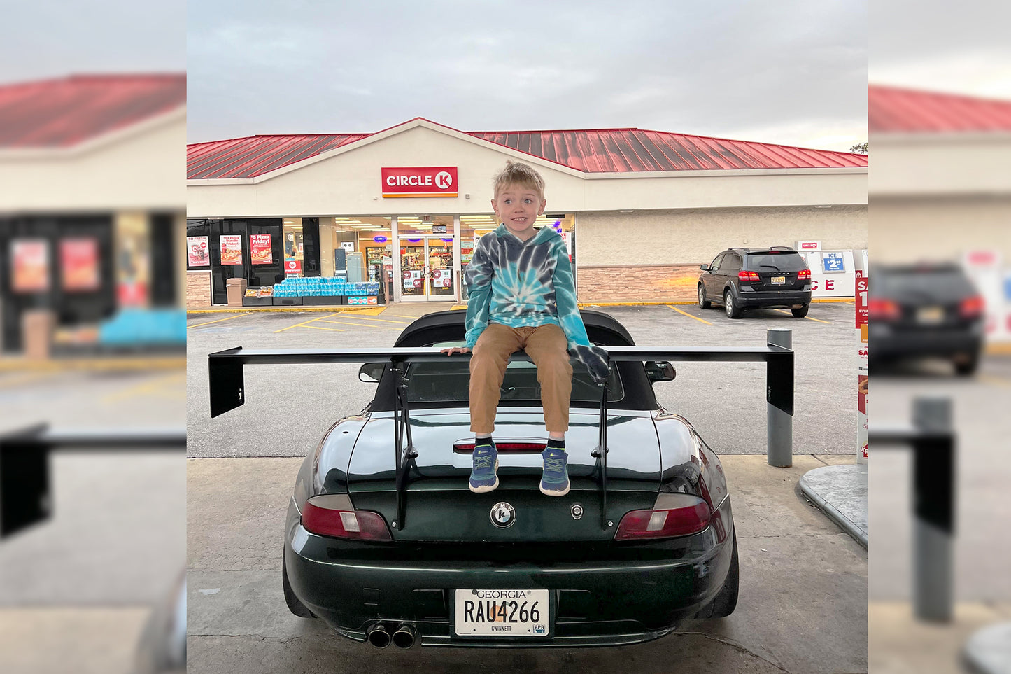 A young boy wearing a tie-dye hoodie and khaki pants is sitting on the 9livesracing BMW Z3 Roadster wing of a black sports car parked in front of a Circle K convenience store. The boy appears happy, and the store's signage and other parked cars are visible in the background.
