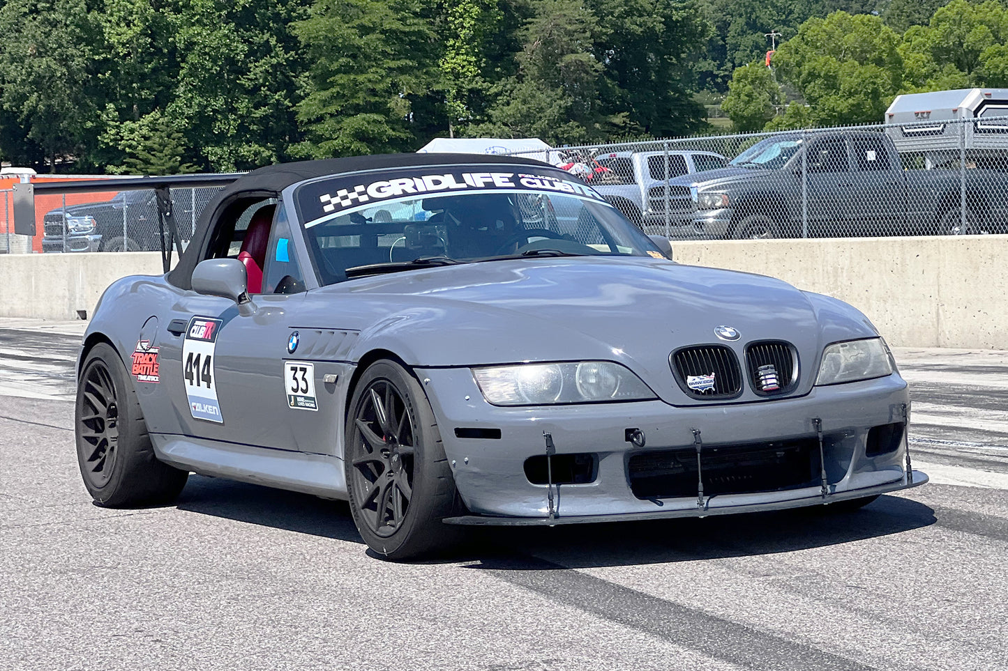 A gray BMW z3 roadster with the number 14 on the driver's door and 'Gridlife' branding on the windshield, sporting 9livesracing wings for improved lift-to-drag ratio and Big Wang GT3 performance modifications, speeds down a track with trees in the background and a barrier separating it from parked vehicles.