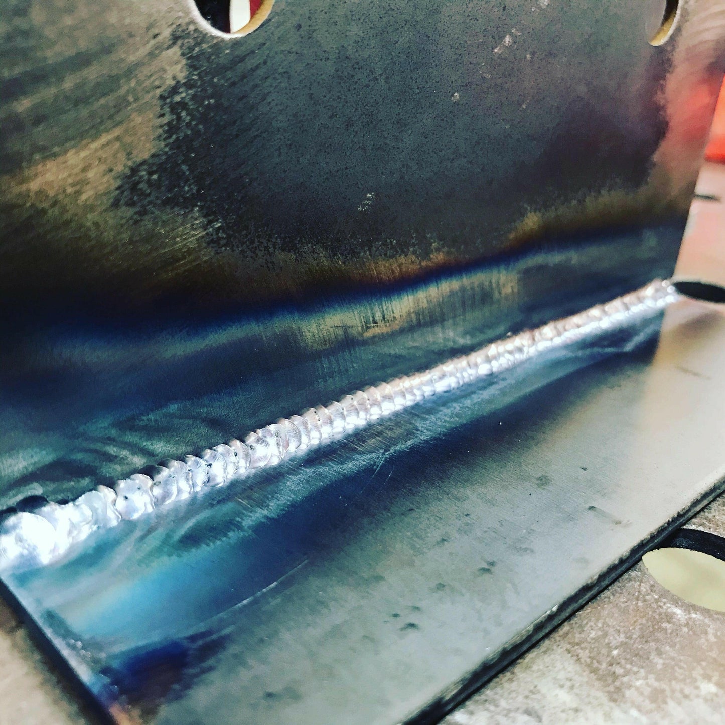 Close-up of a metal piece with a smooth, evenly spaced weld seam in the middle. The surface of the Custom Chassis Mount kit from 9livesracing shows slight discoloration from the heat of welding, varying from blue to brown tones. The background is slightly blurred, highlighting the precision and consistent drive in achieving such craftsmanship.