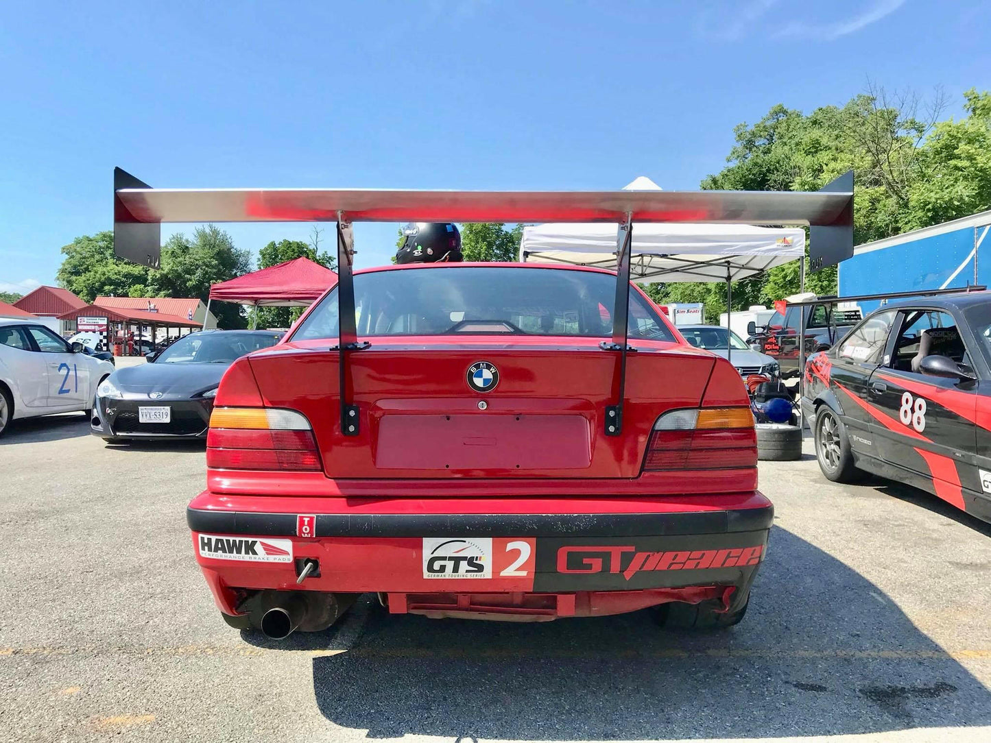 A red race car featuring the 3-Series Big Wang Kit '90-00 E36 by 9livesracing, along with a dual exhaust system, is parked at what appears to be a racetrack. The car sports a GTS class 2 sticker and several other decals. In the background, there is a person wearing a helmet among other race cars and tents.