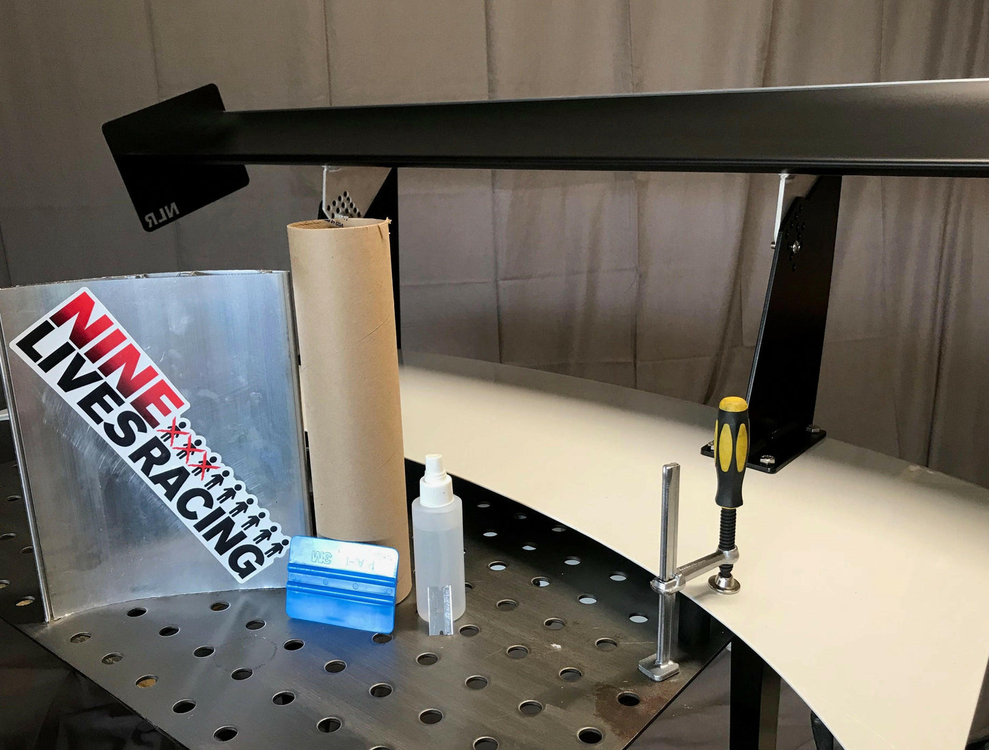 A perforated workbench features a black rear wing, a roll of brown paper, a bottle of hand sanitizer, a blue squeegee tool, and a screwdriver. Nearby hangs a sign displaying the "NINE LIVES RACING" text with its logo. The pre-cut vinyl and car cover for Wang skins- DIY Wang protection by 9livesracing are neatly arranged beside the other tools.
