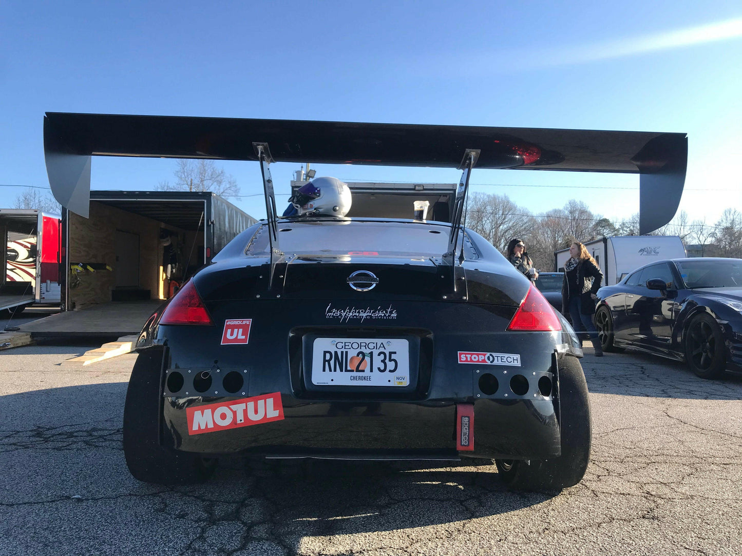 A black sports car with a massive 9livesracing pre-preg carbon wing and various decals, including Motul and StopTech, is parked on a paved area. The license plate reads "RNL7135" from Georgia. A helmet is resting on the rear of the car, and a secondary car is visible nearby.