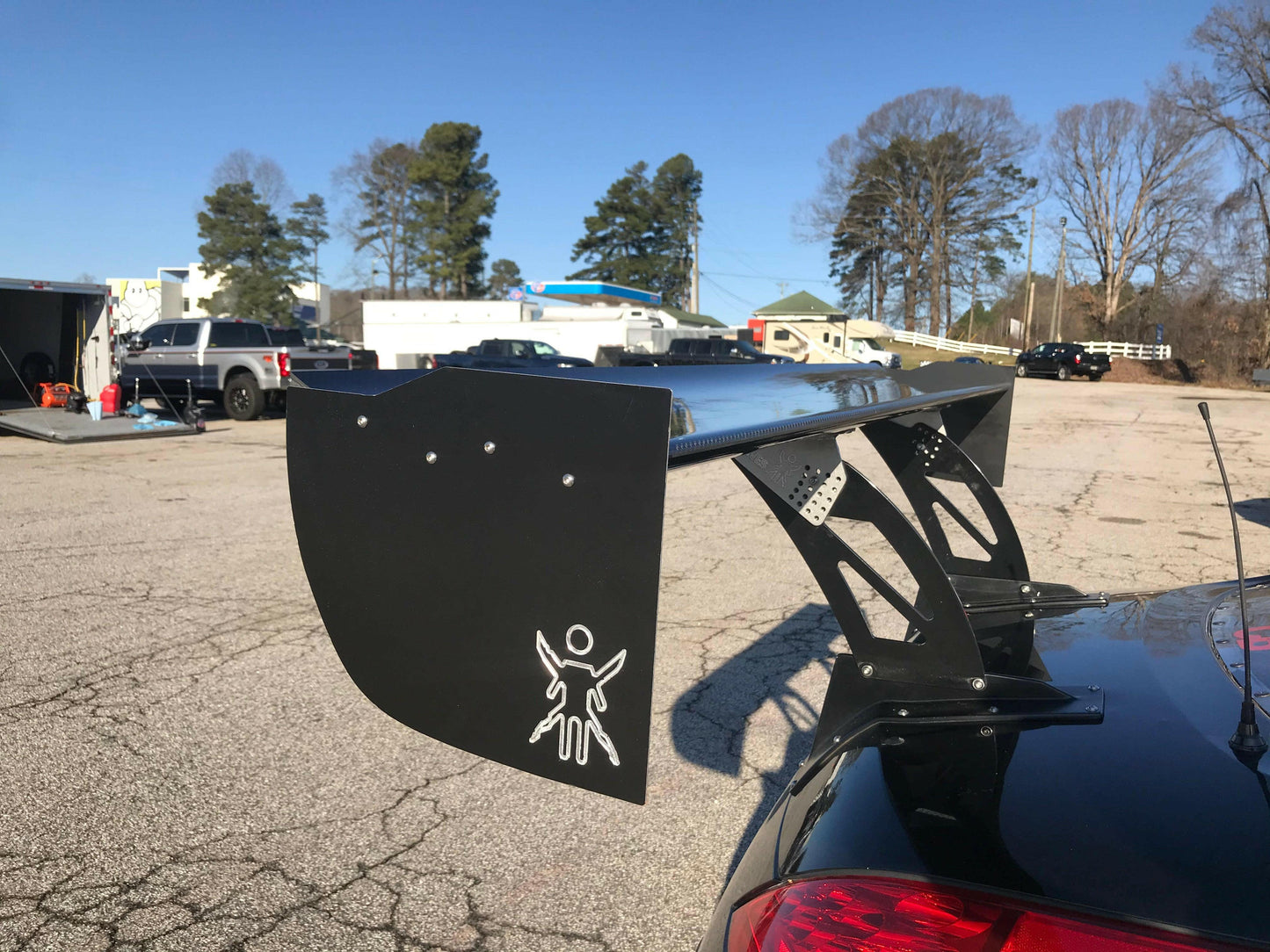 A black sports car, equipped with a large 9livesracing carbon wing, sits in a parking lot. The spoiler, designed to maximize down-force, prominently features the distinctive 9livesracing logo of a figure with spread arms. In the background, other parked vehicles, buildings, and tall trees stand under a clear blue sky.