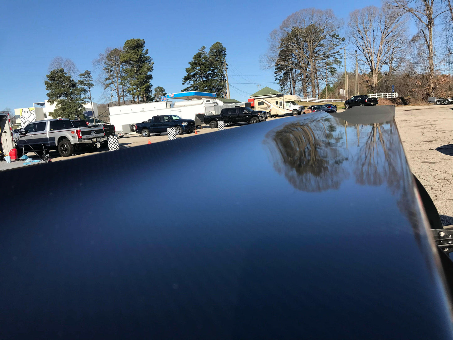 A close-up shot of a sleek, reflective surface, likely a car hood featuring a 9livesracing carbon wing. Trees and parked vehicles are visible in the background under a clear blue sky, capturing a crisp day with bare trees and several black vehicles lined up.