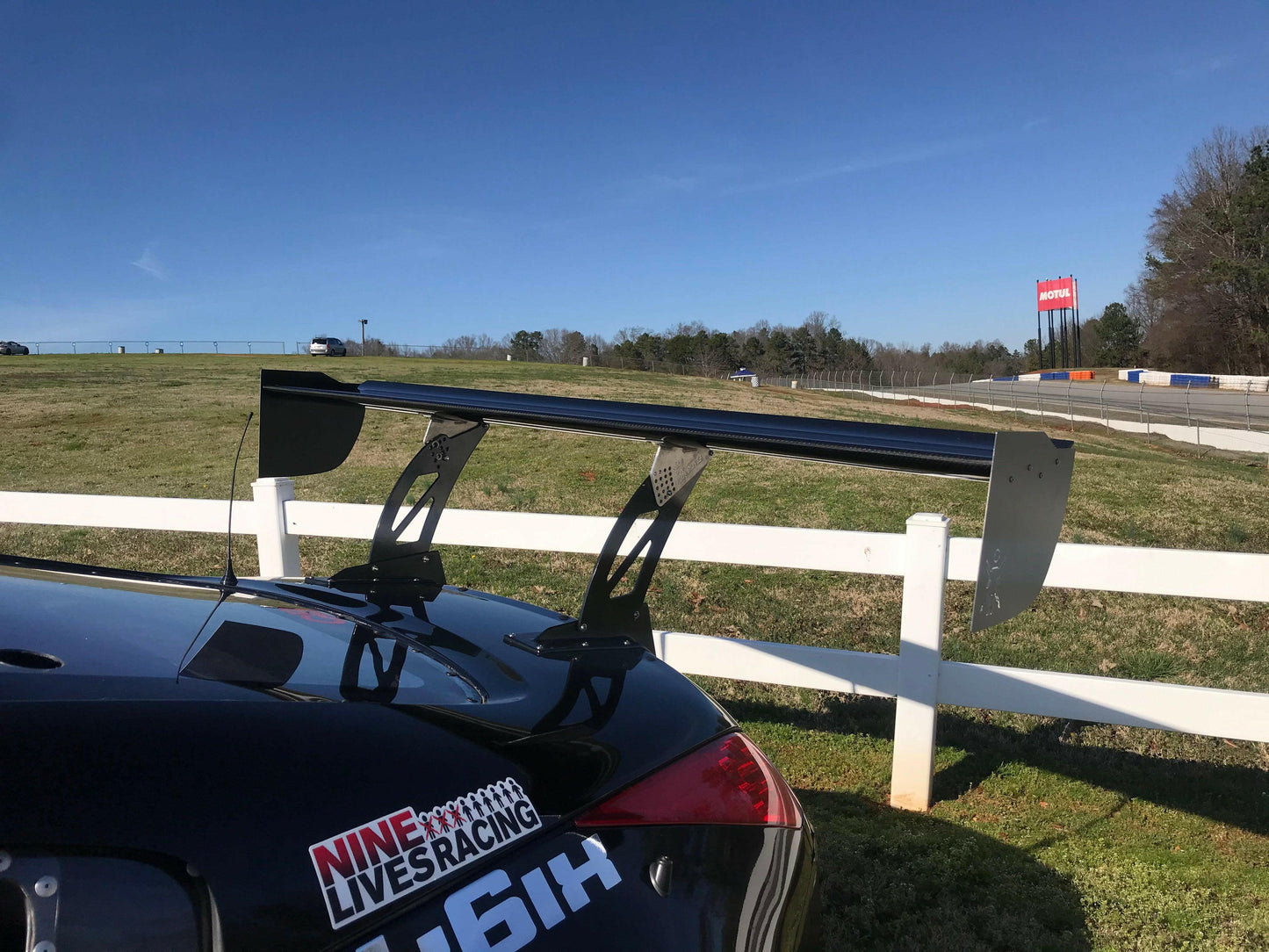 A rear view of a black sports car with a large 9livesracing carbon wing, parked near a white fence. The logo "9livesracing" is visible on the car. A grassy area and a racetrack with barriers are in the background under a clear blue sky.