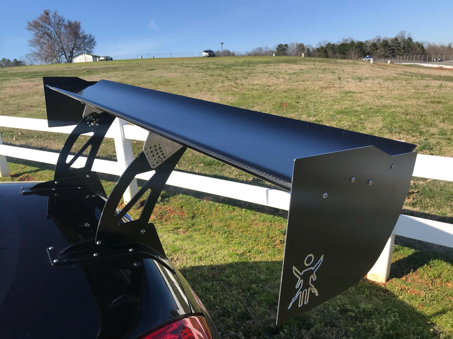 Close-up of a large, black pre-preg 9livesracing carbon wing on a car, set against a rural backdrop with a green field and clear blue sky. The spoiler features the distinct 9livesracing logo with a stylized figure on one side panel. A white wooden fence runs parallel to the car in the background.