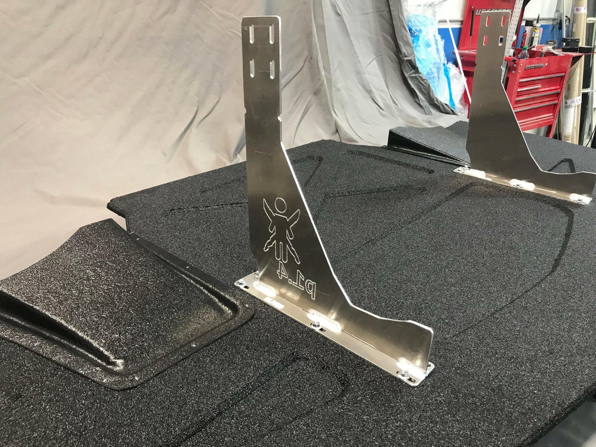 Two metal brackets from the Corvette Medium Downforce Kit ‘97-04 C5 by 9livesracing are installed on a textured black surface. The brackets feature small cutouts, with each one displaying an engraved figure with outstretched arms and the label "D-1." In the background, a workbench with red drawers and various tools can be seen, likely parts of this aerodynamic kit for the C5 Corvette.