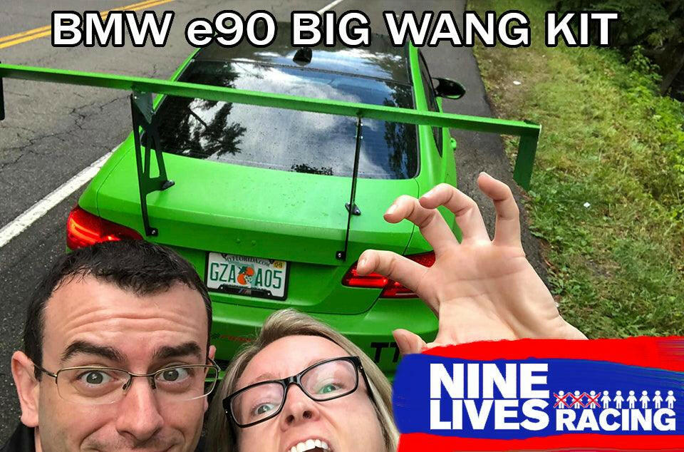 A green BMW 3-Series E90, featuring a 3-Series Big Wang kit ’04-13 E90/E92/E93 with an extruded aluminum wing, is parked on the road. Up front in the photo, two people are smiling and making playful faces. The text "BMW 3-Series BIG WANG KIT" appears at the top, while the "9livesracing" logo is positioned in the lower right corner.