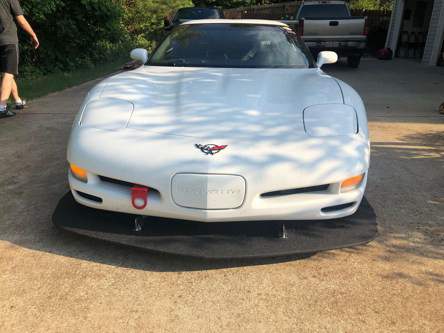 A white C5 Corvette with a 9livesracing Corvette Medium Downforce Kit ‘97-04 C5, featuring a front splitter and a red tow hook, is parked in a driveway. Two people are partially visible in the background near the car. Trees and another vehicle are in the background on a sunny day, highlighting the aerodynamic kit designed for optimal downforce.
