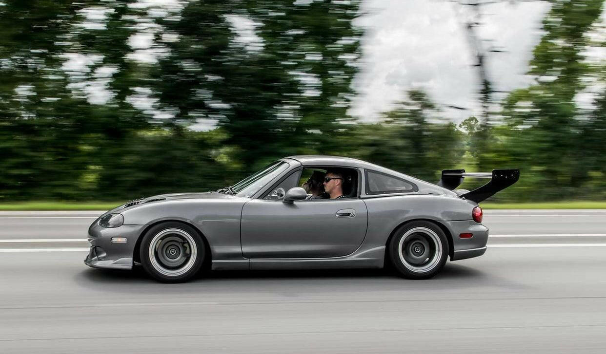 A grey sports car, equipped with a large rear spoiler from the Miata V2 Street Wang '90-05 NA/NB by 9livesracing, is seen speeding down a road. The driver, donning sunglasses and black clothing, can be seen through the open window. The background features trees and a cloudy sky, with motion blur highlighting the fast movement.