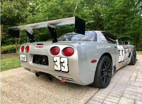 A silver Corvette race car with the number 33 displayed on its sides and back is parked on a paved driveway. The car boasts 9livesracing's Corvette C5/C6 Mounts - Pylons Only for added stability, along with a large rear spoiler, black wheels, and various decals. Trees and greenery are visible in the background.