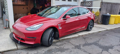 A sleek red Tesla Model 3, equipped with the 9livesracing Big Wang GT3 performance aluminum wings, is parked halfway onto the curb in a residential neighborhood. The car is positioned outside a garage filled with various tools and items inside. To the right are three garbage bins: one green, one yellow, and one black.