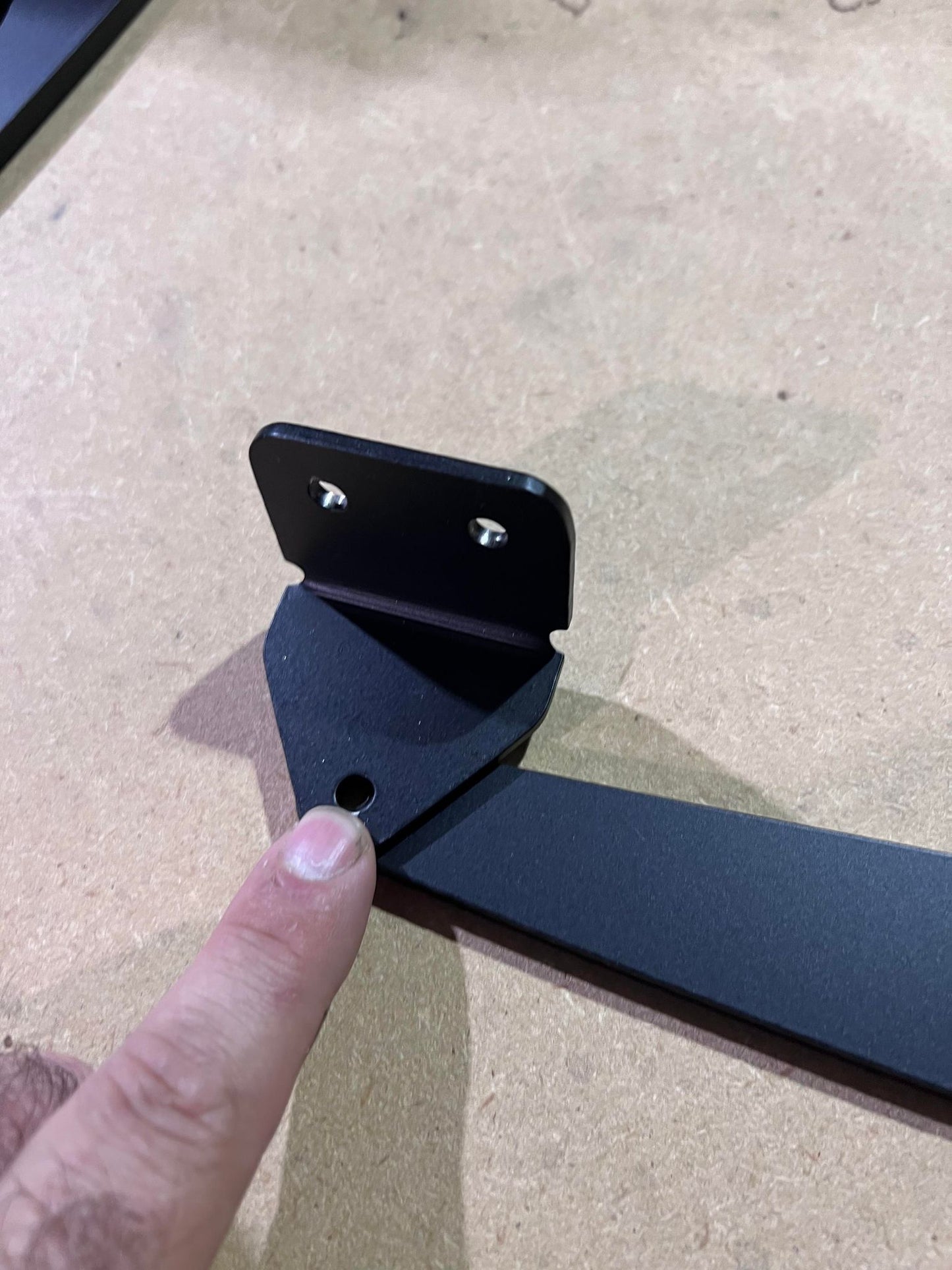 A finger pointing at the hole in a black metal bracket on a tabletop, suggesting alignment or assembly for the adjustable mount angle of the Universal Hatchback Wing Pylons by 9livesracing.