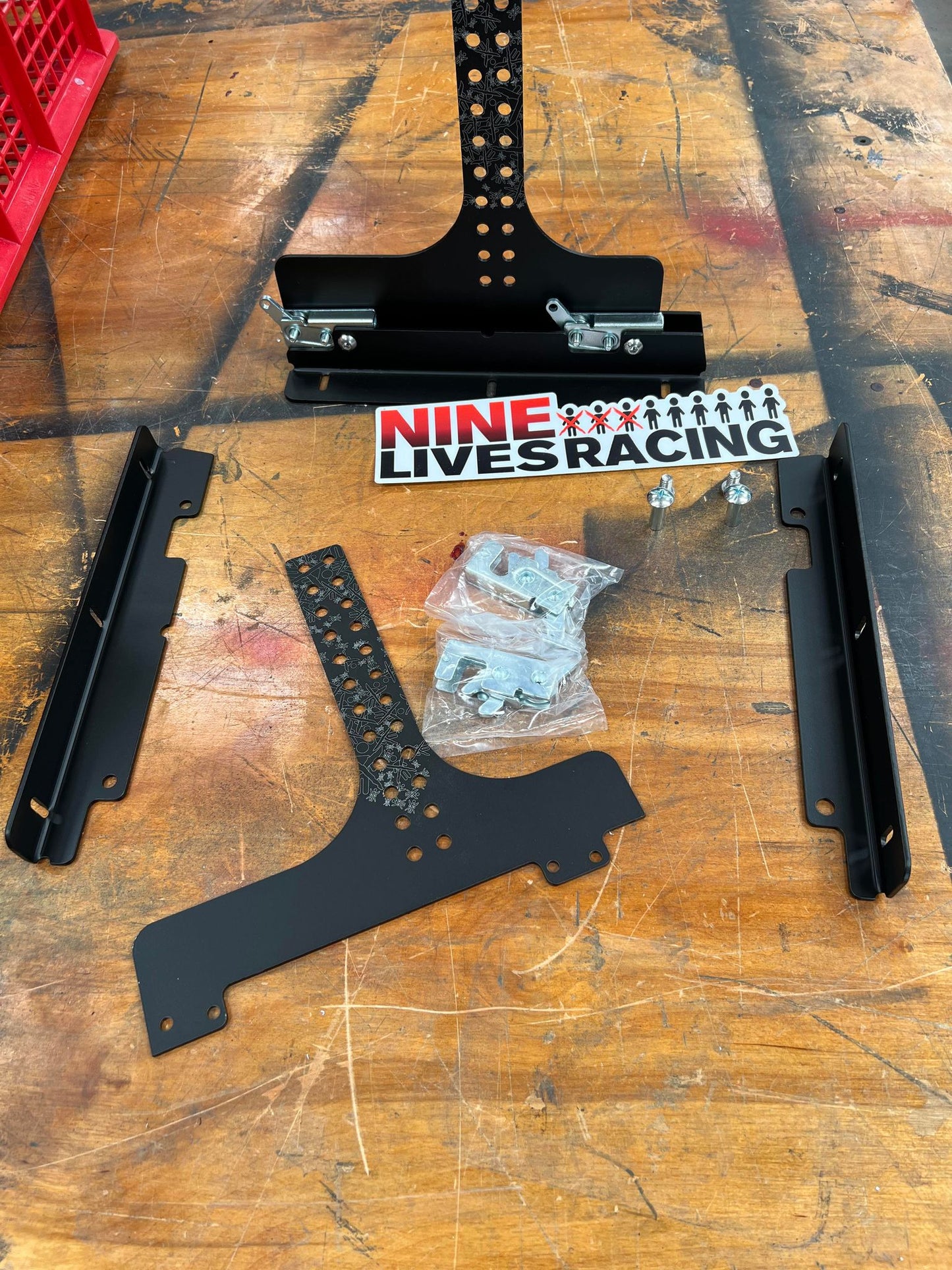 The image displays a disassembled Nine Lives Racing Chassis Mount Splitter Quick Release Kit laid out on a wooden table. Visible components include black metal brackets, splitter mounts, a perforated metal strip, screws in a plastic pouch, and a Nine Lives Racing logo sticker. A red container is partially visible in the corner.