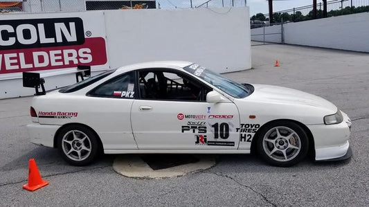 A white racing car with the number 10 and various sponsor decals, including "Honda Racing" and "Toyo Tires," is parked on an asphalt surface beside orange cones. The car, equipped with a 9livesracing Integra Wang kit '94-01 for maximum downforce adjustments, has a large rear spoiler and a "PKZ" decal on its rear window. A "Lincoln Welders" sign is in the background.