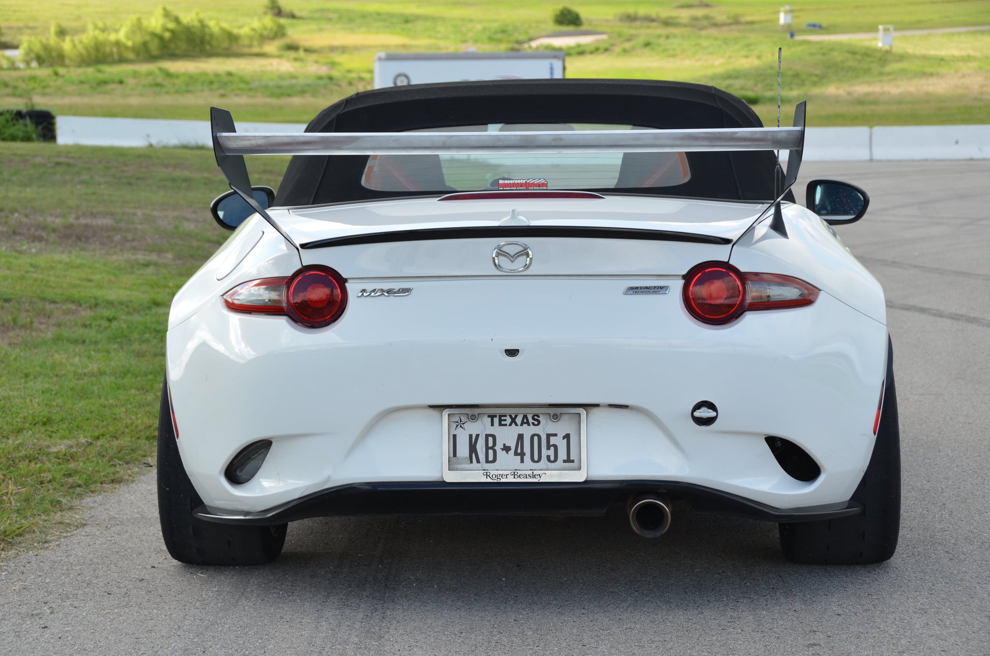 A white Mazda Miata Street Wang 2016+ ND, equipped with a low downforce package and raised spoiler from 9livesracing, is parked on a paved surface against a grassy background. The Texas license plate reads "K8*4051," and the car's top is down, revealing part of the interior that any track day lover would admire.