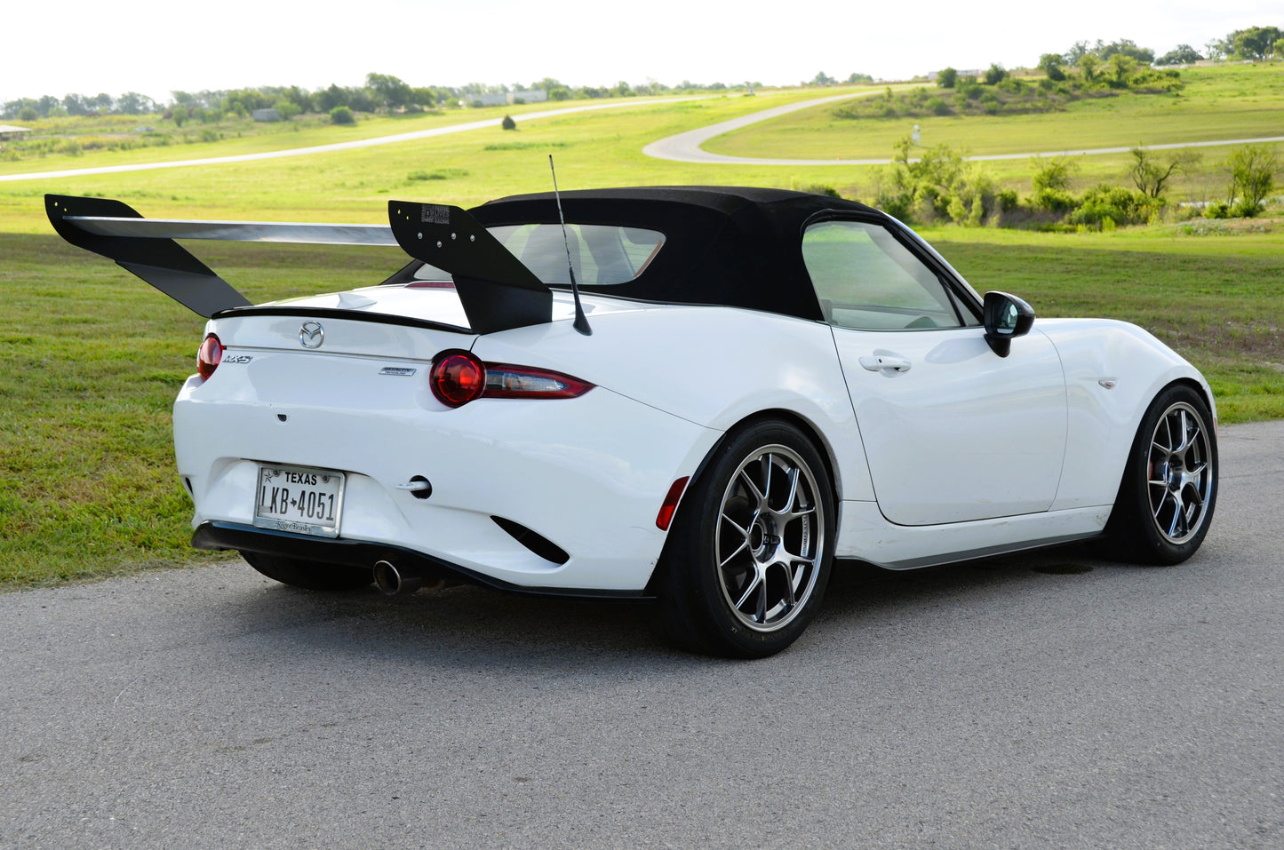 A white Miata Street Wang 2016+ ND by 9livesracing, equipped with a black soft top, alloy wheels, and a large rear spoiler giving off Track day lover vibes, is parked on a paved road. The car flaunts sleek lines and displays a visible Texas license plate. In the background lies an open green landscape with a winding path and distant trees.