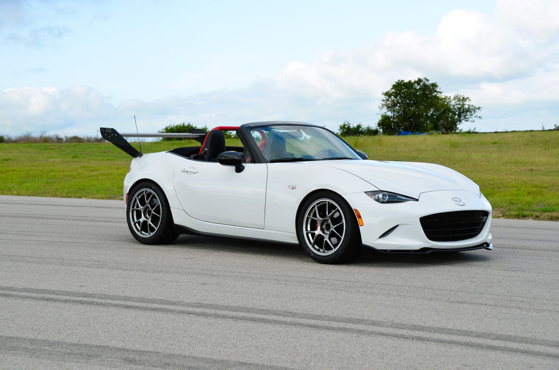 A white Mazda MX-5 Miata convertible, favored by track day enthusiasts, is parked on an empty road. The car boasts a sleek, sporty design with a prominent black rear spoiler from the Miata Street Wang 2016+ ND kit by 9livesracing and stylish modern alloy wheels. The background features a grassy field and a partly cloudy sky.