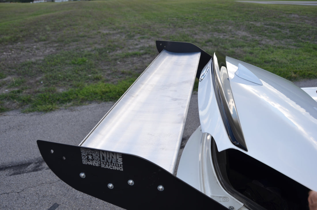 Close-up of a white sports car’s rear spoiler from the Miata Street Wang 2016+ ND kit, featuring a sleek metallic design with a logo on the end plate that reads "9livesracing." The background shows a grassy area next to a paved road or parking lot. Ideal for any track day lover.