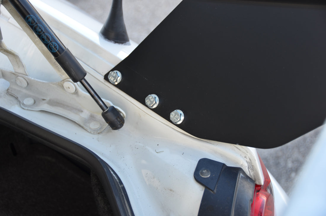 Close-up of a white car's trunk lid showcasing the hydraulic strut and mounting bolts of the 9livesracing Miata Street Wang 2016+ ND. The image focuses on the attachment point, featuring visible rivets and a black metal strut supporting the spoiler—perfect for any track day lover's low downforce package needs.