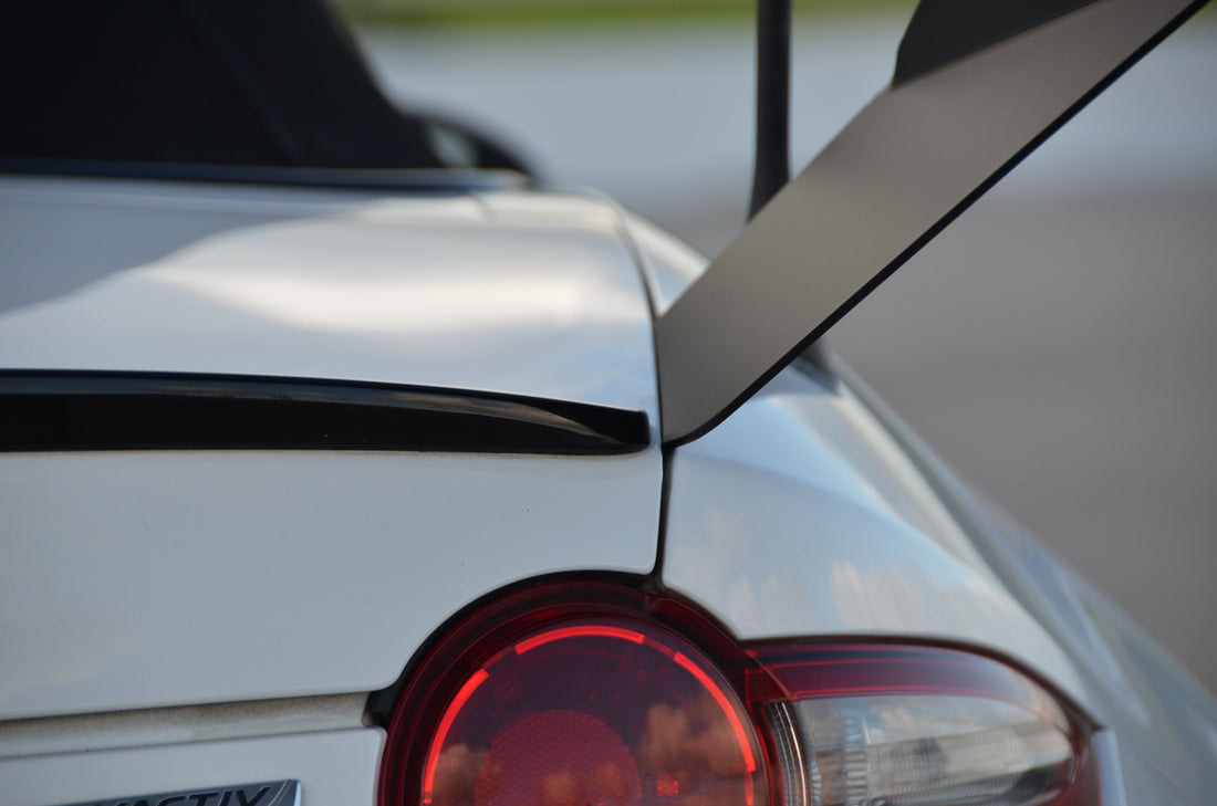 Close-up of the rear of a white Miata Street Wang 2016+ ND by 9livesracing, featuring the red tail light and a prominent rear spoiler enhanced by a low downforce package. The image highlights the sleek design and aerodynamic features, perfect for any track day lover. The background is blurred, drawing attention to the car's details.