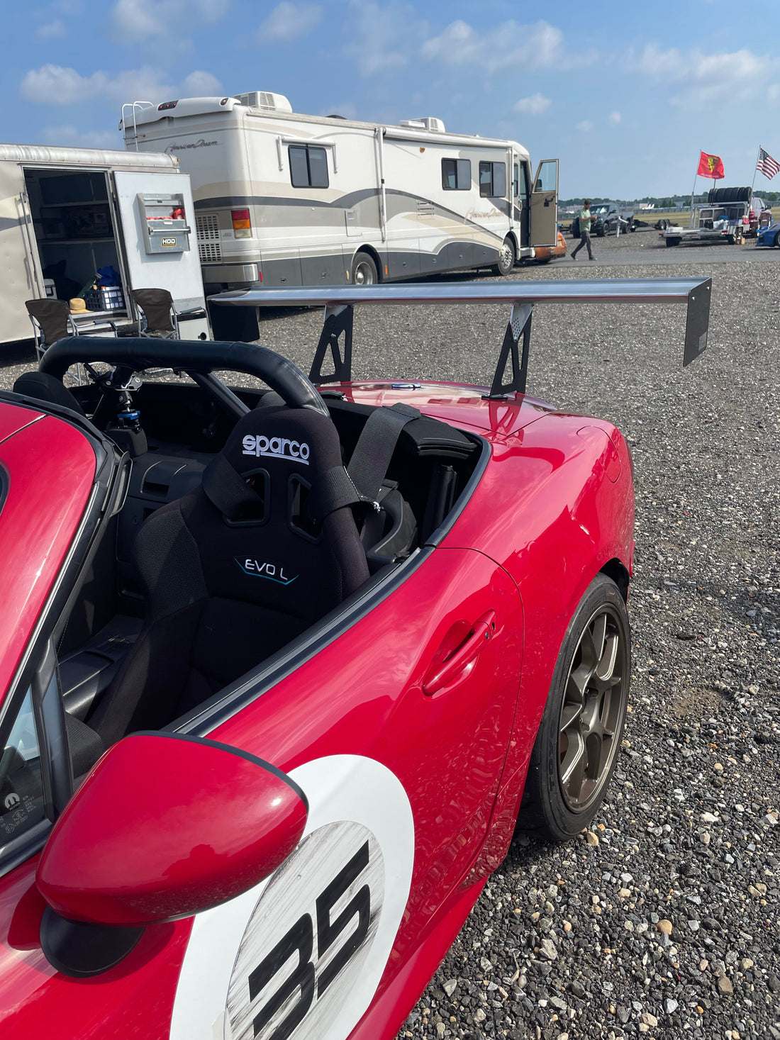 A Fiat 124 Spider equipped with a Fiat 124 Spider Big Wang Kit 2016+ by 9livesracing and wearing the number 35 on its door is parked on a gravel surface near trailers and an RV. It features a Big Wang GT3 rear wing for optimal down-force and a Sparco Evo L racing seat. The background shows a cloudy sky and a few flags.