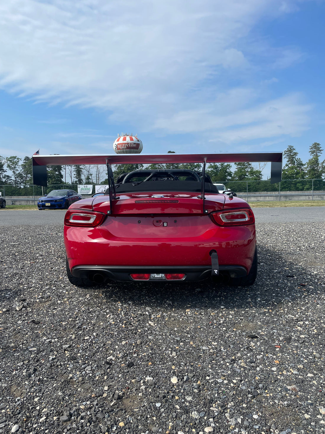 A red Fiat 124 Spider featuring the 9livesracing Big Wang Kit GT3 rear wing and a racing helmet resting on top, parked on a gravel surface. A blue car is closely parked nearby with fencing in the background. Trees and a partly cloudy sky are visible in the distance.