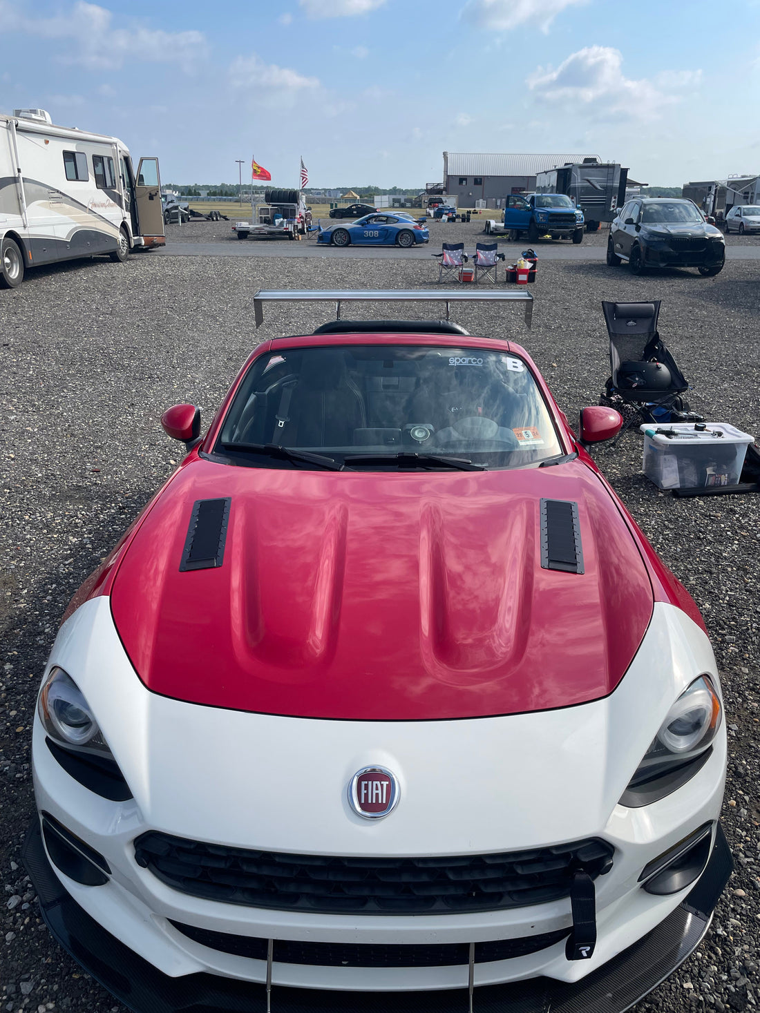 A red and white Fiat 124 Spider Big Wang Kit 2016+ by 9livesracing, featuring two black hood vents, is parked in a gravel lot at a racetrack. Behind it are other vehicles, including RVs and more cars, along with a building structure under a partly cloudy sky. The car proudly features an airfoils down-force design to enhance its performance.