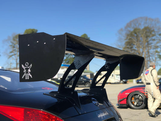 Close-up of a black car with End Plates for Carbon Wáng from 9livesracing and a large rear spoiler featuring a white angel logo. A red and black car is parked beside it in the background, and a person in light-colored racing gear is walking nearby. Trees and buildings are visible in the background.