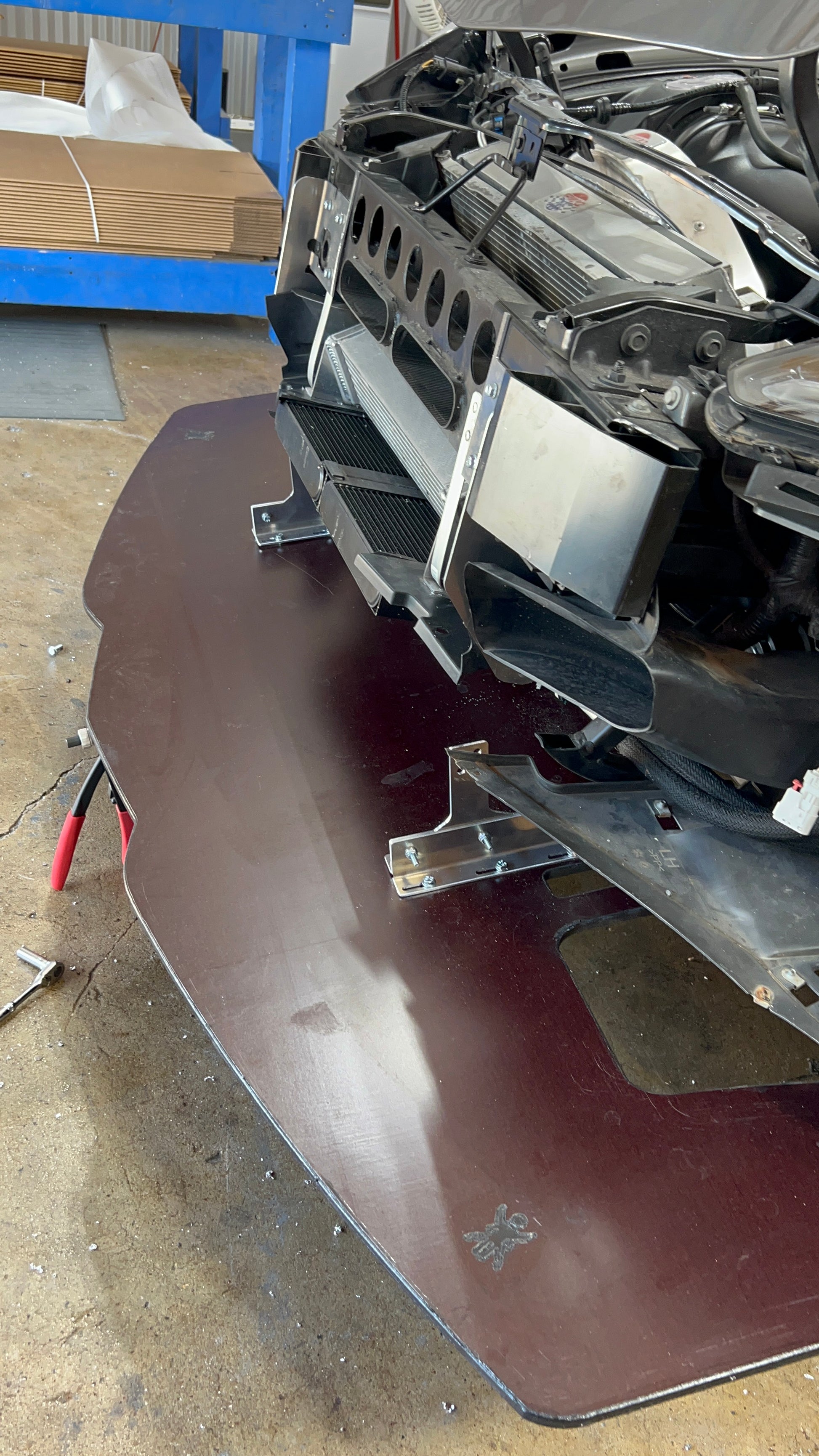 Close-up of a car's front end under repair, showcasing the exposed mechanical parts and an angled front splitter. The image hints at the installation of a Corvette splitter kit C7 with Sturdy Boii mounts from 9livesracing. Tools and components are scattered on the garage floor. Blue-painted shelving with various items is visible in the background.