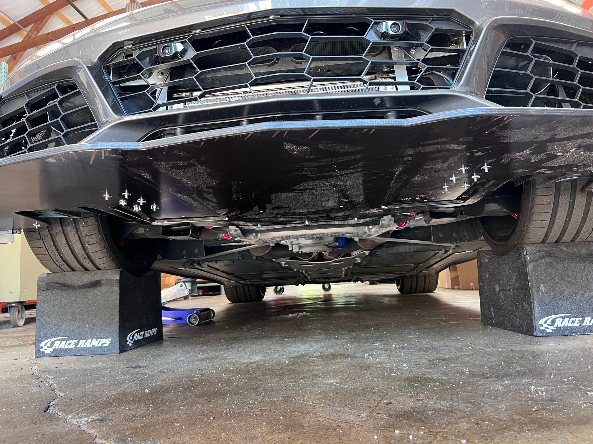 A close-up of a car's undercarriage, elevated on Race Ramps. The black front bumper and grille are slightly visible, showcasing a Corvette splitter kit C7 with Sturdy Boii mounts from 9livesracing for added downforce. The car is in a garage setting with visible details of the underside including various mechanical parts and components.