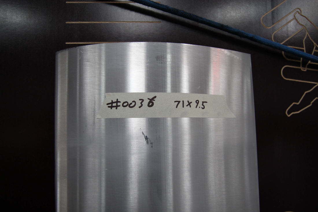 A close-up view of a cylindrical metal object with a piece of masking tape on which "#0036 71x9.5" is written. The smooth, reflective surface sits against a background showing part of a black surface with a blue line and a partial graphic pattern, possibly prepared for shipping. This is from the Nine Lives Racing Buy Aluminum Motorsport Wing Kits collection, specifically the "71" wang 9.5" spacing - #0036.