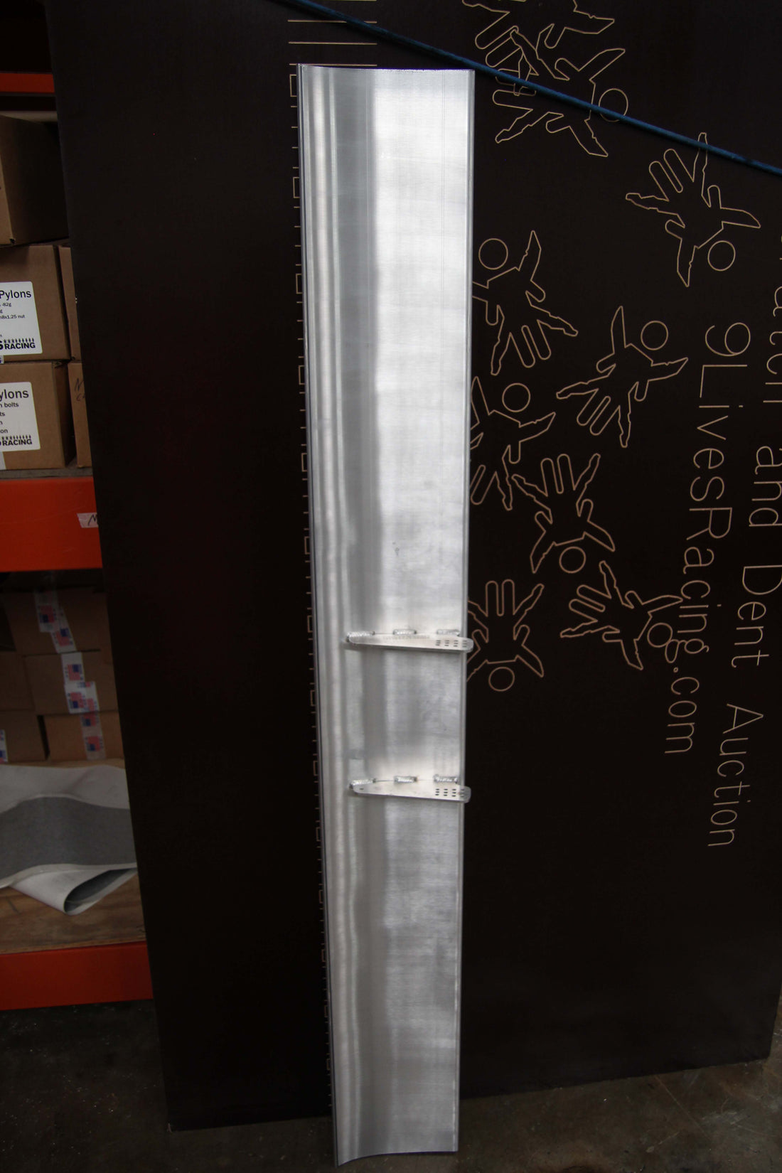 A tall, silver metallic 71" wang 9.5" spacing - #0036 vertical panel is positioned against a dark background adorned with printed designs and text. Two small brackets are attached to the panel. On the left side of the image, shelves and boxes are partially visible, ideal for organizing shipping materials with their 9.5" spacing. This product is from Nine Lives Racing Buy Aluminum Motorsport Wing Kits.