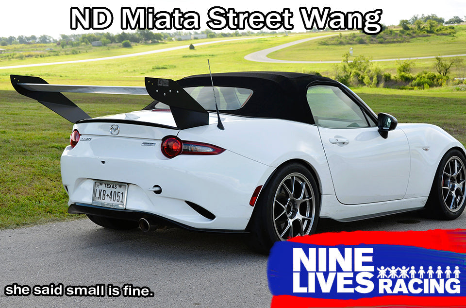 A white Mazda Miata convertible featuring a large rear wing from the Miata Street Wang 2016+ ND kit is parked on a grassy area overlooking a scenic landscape. The image displays the text "ND Miata Street Wang" at the top, accompanied by the 9livesracing logo and the phrase "she said small is fine." at the bottom.