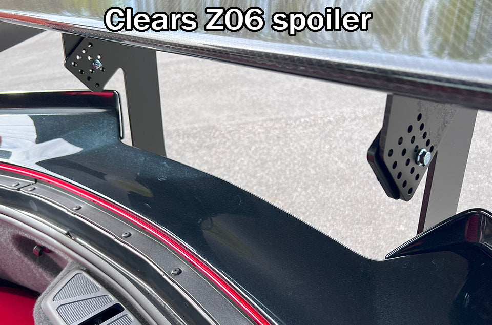 Close-up view of a rear Corvette Big Wang Chassis Mount '2020 C8 spoiler attached to a car, showcasing the mounting brackets and structure. The car's trunk area is visible beneath the spoiler, revealing part of the interior and detailing. Text at the top reads, "Clears Big Wang Chassis Mount," highlighting its excellent lift-to-drag ratio for optimal down-force. This premium spoiler by 9livesracing provides superior aerodynamics for your Corvette.