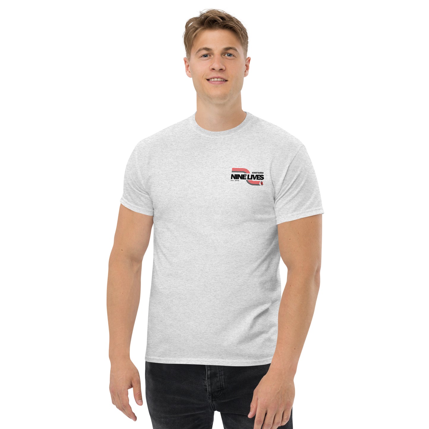 A smiling man with short hair wears a light gray Men's Soda Can Wang t-shirt from Nine Lives Racing, featuring the text "NINE LIVES" on the chest in black and red. He is standing against a plain white background, dressed in black pants and embodying a sleek, 100% cotton look perfect for layered streetwear outfits.