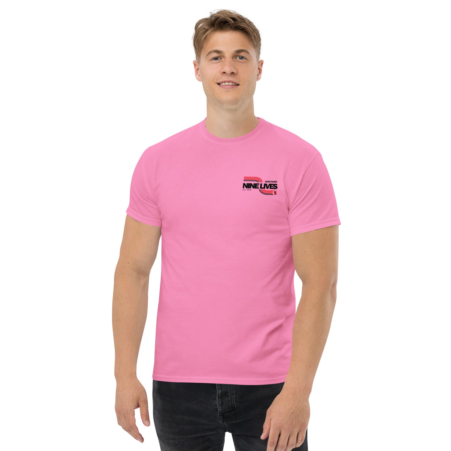 A smiling man with short hair is wearing a pink, 100% cotton Men's Soda Can Wang t-shirt from Nine Lives Racing. The shirt features a small logo on the left chest that reads "NINE LIVES" in black text. He is standing against a plain white background and pairing his tee with black pants, completing his understated layered streetwear outfit.