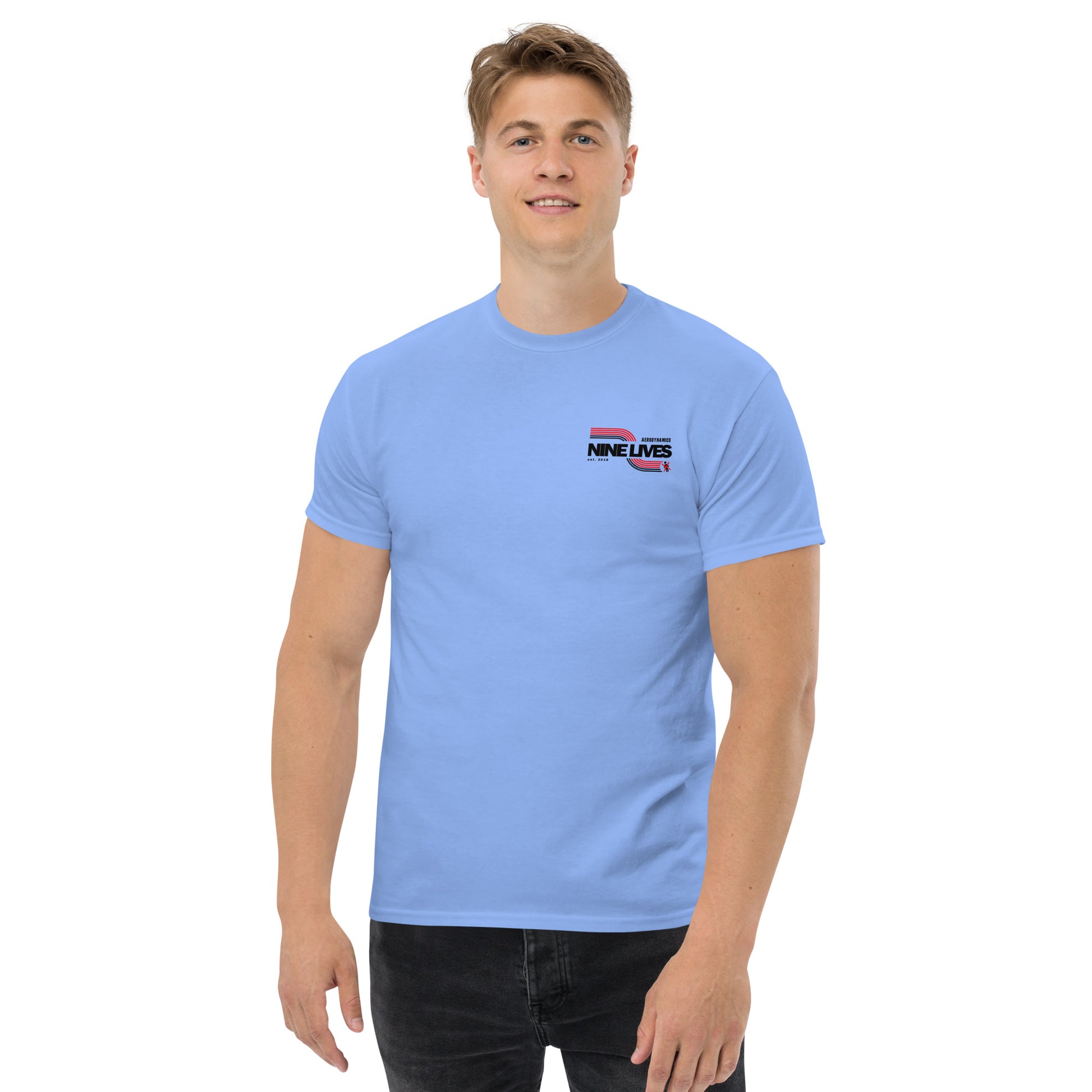 A young man with light brown hair stands against a plain white background, wearing a light blue Men's Soda Can Wang t-shirt from Nine Lives Racing. The 100% cotton shirt pairs perfectly with his dark jeans, making it ideal for layered streetwear outfits. He smiles with his arms relaxed by his sides.