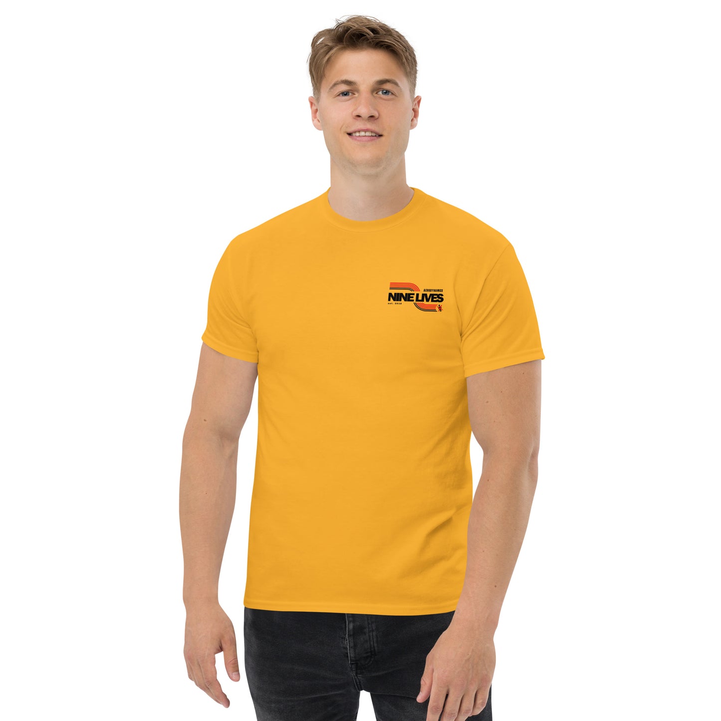 A man with short blond hair stands smiling, wearing a bright yellow Men's Soda Can Wang t-shirt from Nine Lives Racing and black jeans. Made of 100% cotton, the classic tee features a small logo on the left chest that reads "Nine Lives" with a striped design above the text. The background is plain white.