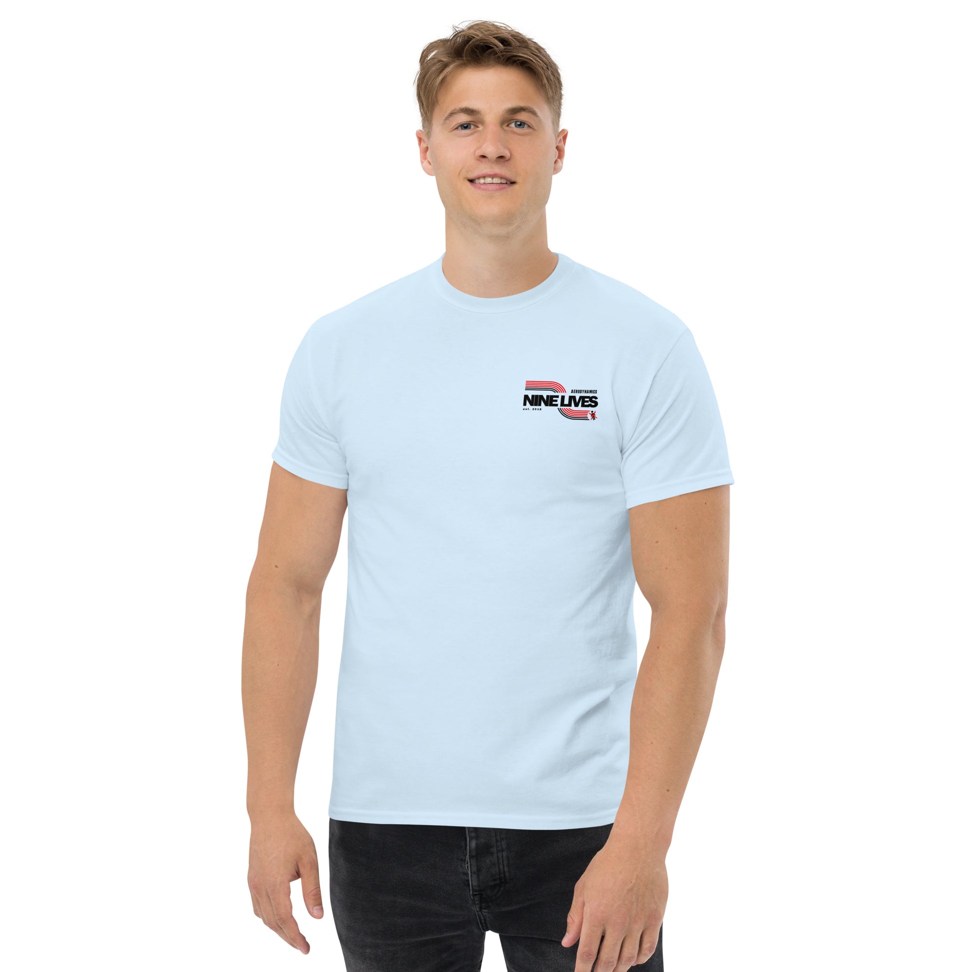 A person with short hair is wearing a light blue Men's Soda Can Wang t-shirt from Nine Lives Racing, made from 100% cotton. The tee features the text "NINE LIVES" and a small graphic on the upper left chest area. They are also wearing black pants, standing against a plain white background, and smiling slightly.