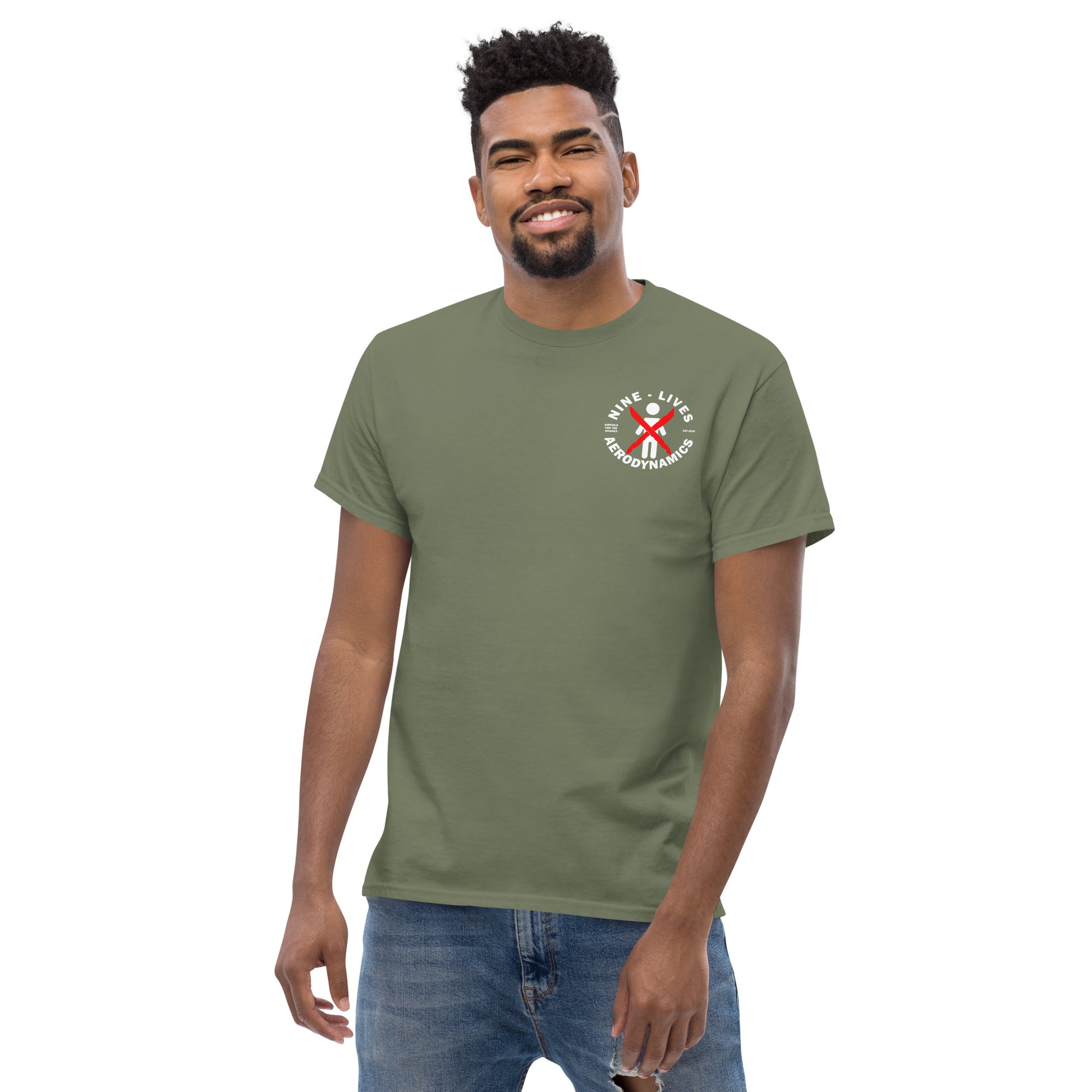 A man wearing an Aerodynamics Is Witchcraft T-shirt (ouija) by Nine Lives Racing, crafted from 100% cotton and featuring a green color with a logo displaying a red ribbon and text that reads "SAVE LIVES CHARGEMAN 2019" on the left chest. He has short dark hair, a beard, and is smiling while also donning blue jeans—perfect for streetwear outfits.