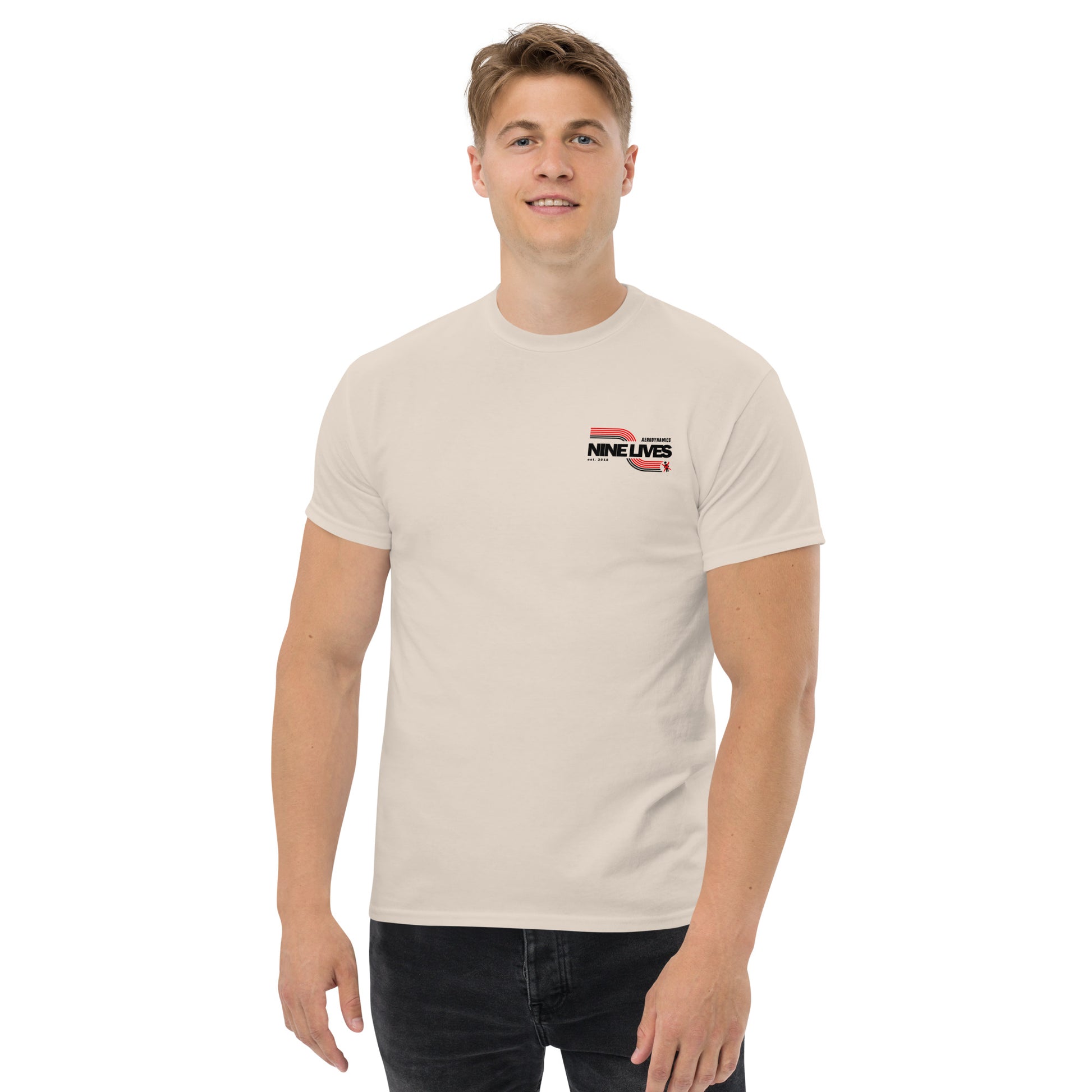 A smiling man stands against a white background, wearing a beige Men's Soda Can Wang t-shirt by Nine Lives Racing that has the text "NINE LIVES" with a small graphic on the left side of his chest. Made from 100% cotton, it’s perfect for layered streetwear outfits. He has short, light brown hair and is also wearing dark pants.