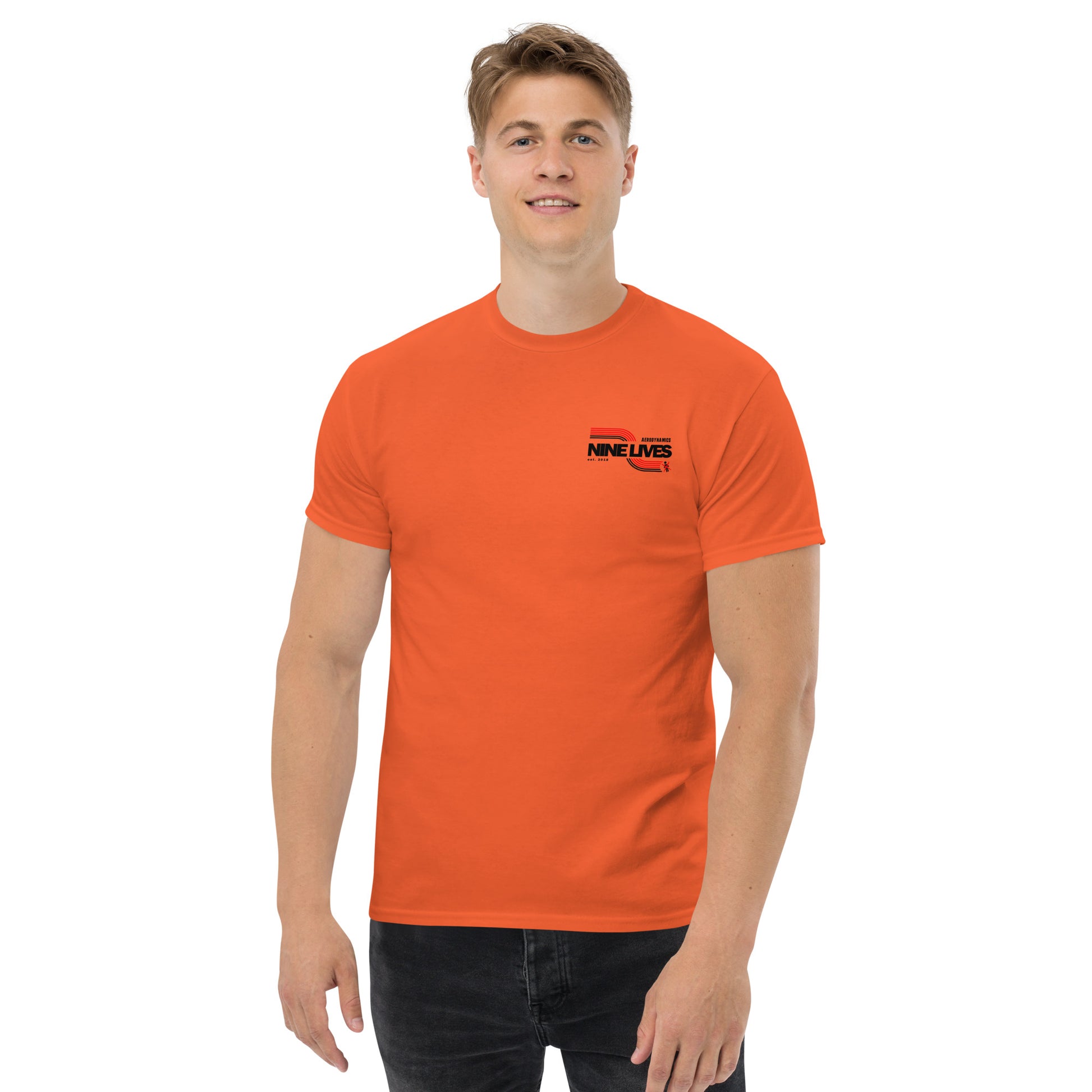 A person with short hair is standing and smiling. They are wearing an orange Men's Soda Can Wang t-shirt from Nine Lives Racing made of 100% cotton, featuring the text "NINE LIVES" and a small logo on the left chest, along with black pants. The background is plain white.