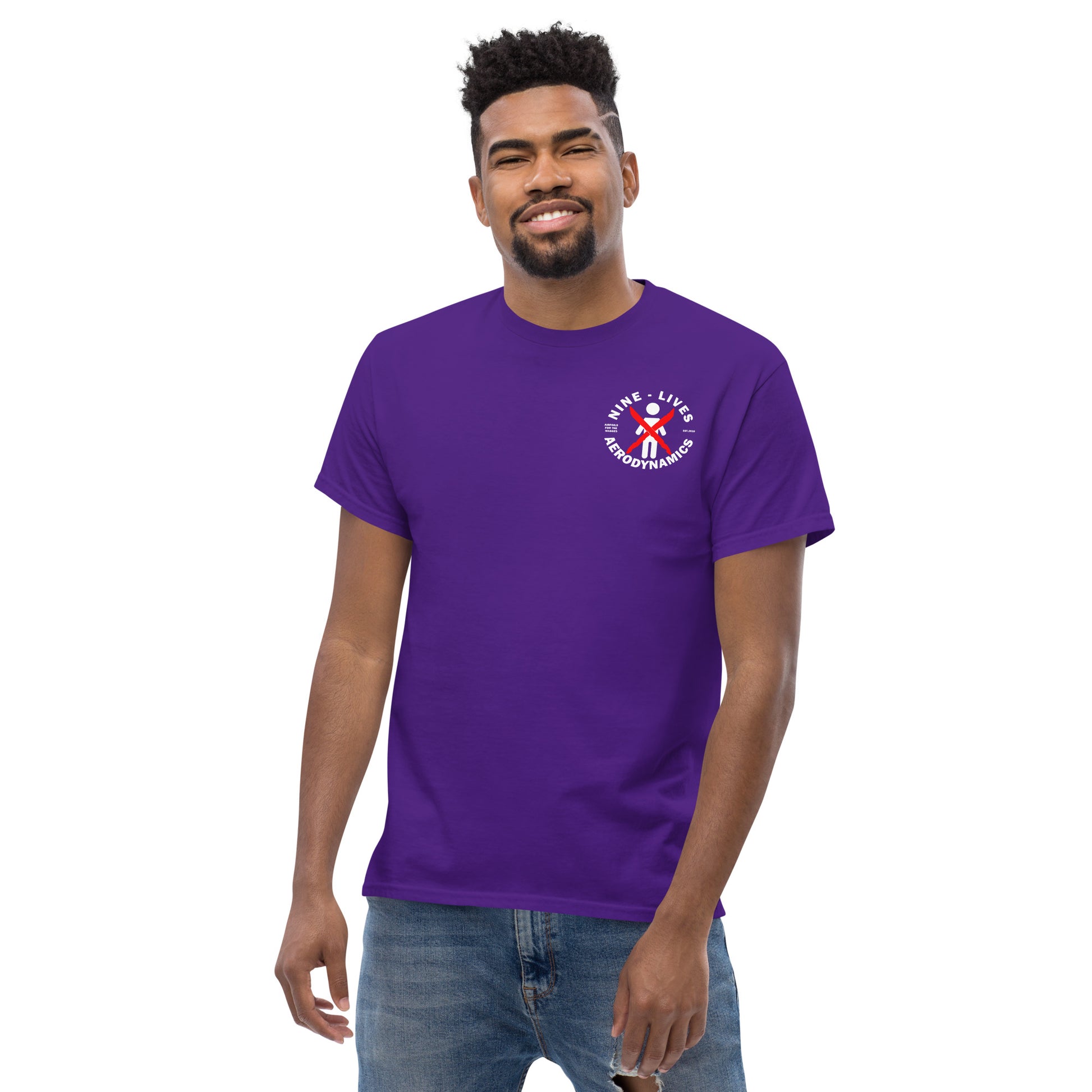 A man with a beard stands against a white background, wearing a 100% cotton, purple "Aerodynamics Is Witchcraft" Men's classic tee from Nine Lives Racing and blue jeans. The T-shirt, part of trendy streetwear outfits, features a small circular graphic on the left chest displaying the words "SAVE LIVES" encircling a medical symbol with a red, white, and blue ribbon.