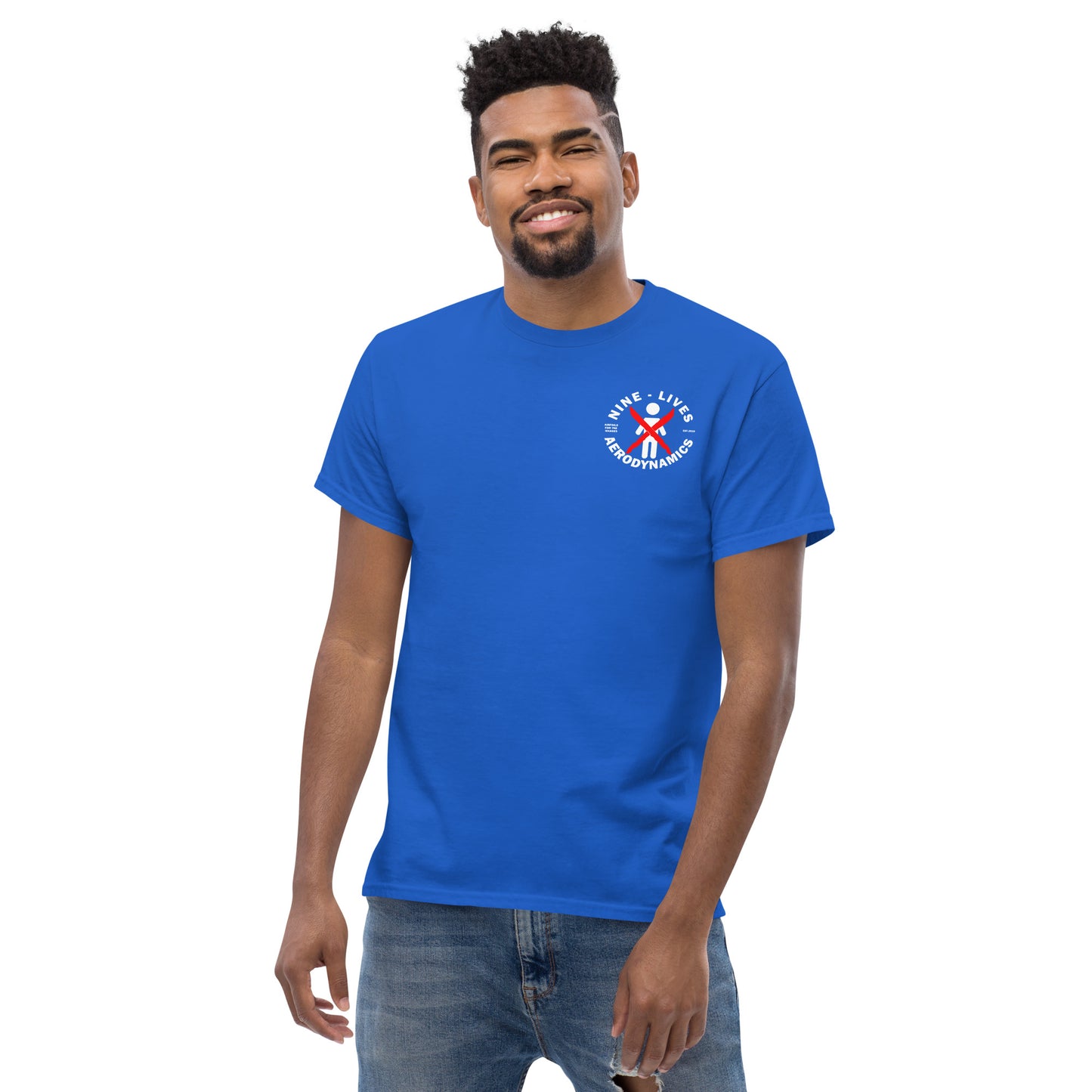 A person with short, curly hair and a beard is wearing a bright blue "Aerodynamics Is Witchcraft" T-shirt made of 100% cotton from Nine Lives Racing. The tee features a small logo on the left chest area with a ribbon in red and blue colors bearing the text "Nine Lives" and "Bloodhound." The person is smiling.