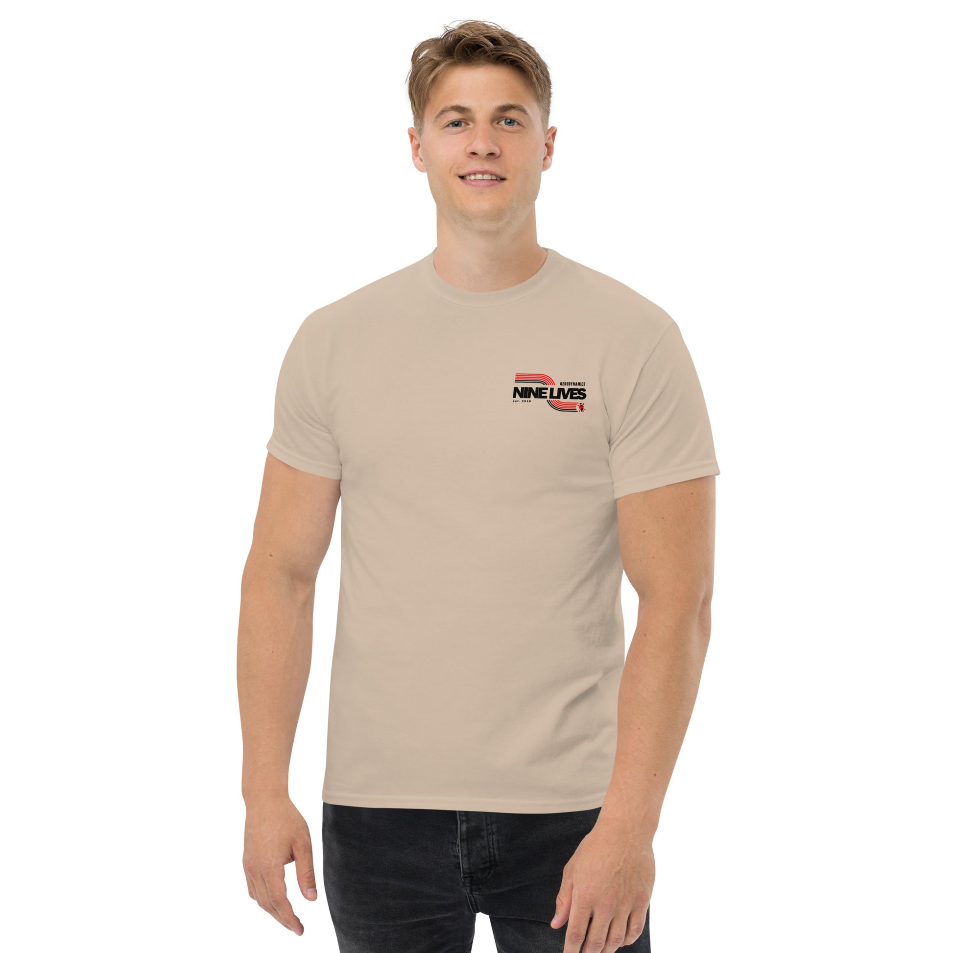 A man with short blonde hair is standing and smiling. He is wearing a beige Men's Soda Can Wang t-shirt from Nine Lives Racing, made from 100% cotton, featuring a small "NINE LIVES" logo on the left side of his chest and black pants. The background is white.