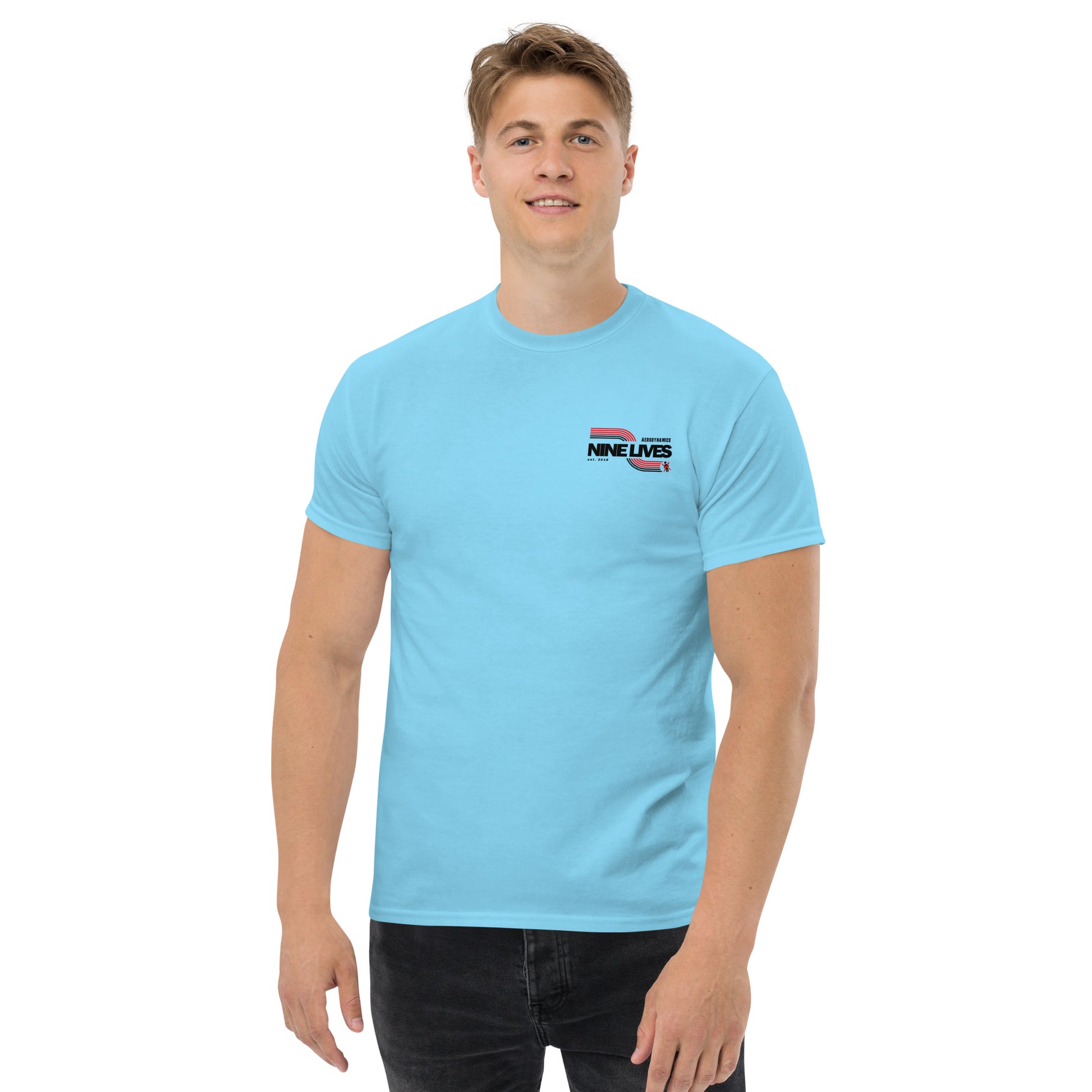 A smiling person with short blond hair is wearing a light blue Men's Soda Can Wang t-shirt by Nine Lives Racing, featuring "NINE LIVES" printed in the top left corner. The 100% cotton T-shirt pairs perfectly with the black pants, creating a versatile look against the plain white background.